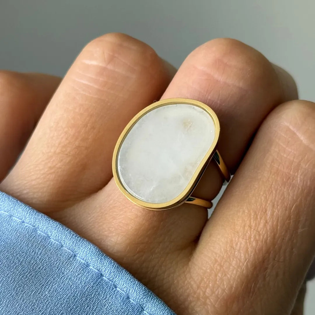 Une bague dorée avec une grande pierre blanche translucide de forme libre, portée sur un doigt à côté d'une manche de chemise