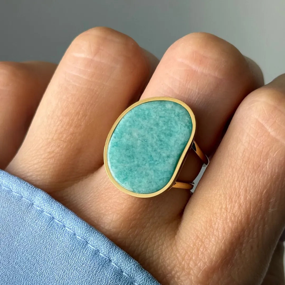 Bague dorée à large pierre ovale irrégulière bleu-vert marbrée, portée au doigt.