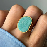 Bague dorée à large pierre ovale irrégulière bleu-vert marbrée, portée au doigt.