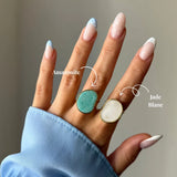 Deux bagues dorées à pierre naturelle irrégulière portées sur une main. L'une est une amazonite verte, l'autre un jade blanc.