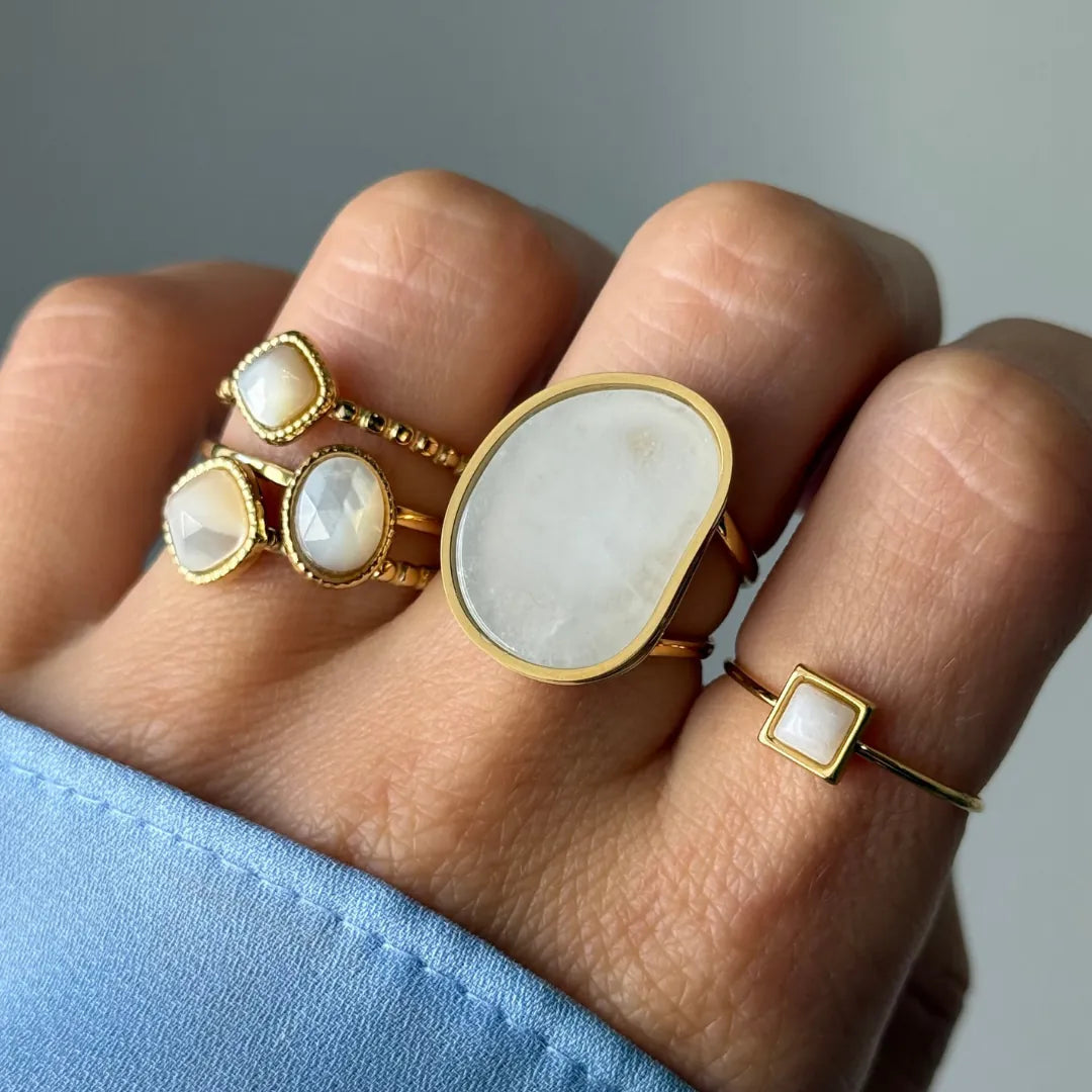 Main portant cinq bagues dorées avec des pierres blanches opalescentes. Une grande ovale, une petite carrée, et trois de