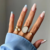 Main portant cinq bagues dorées avec des pierres en nacre blanches. Les ongles sont décorés d'une manucure française bleue à