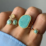 Main portant cinq bagues dorées. Une grande pierre turquoise irrégulière, deux ovales vertes, une carrée crème, et une petite