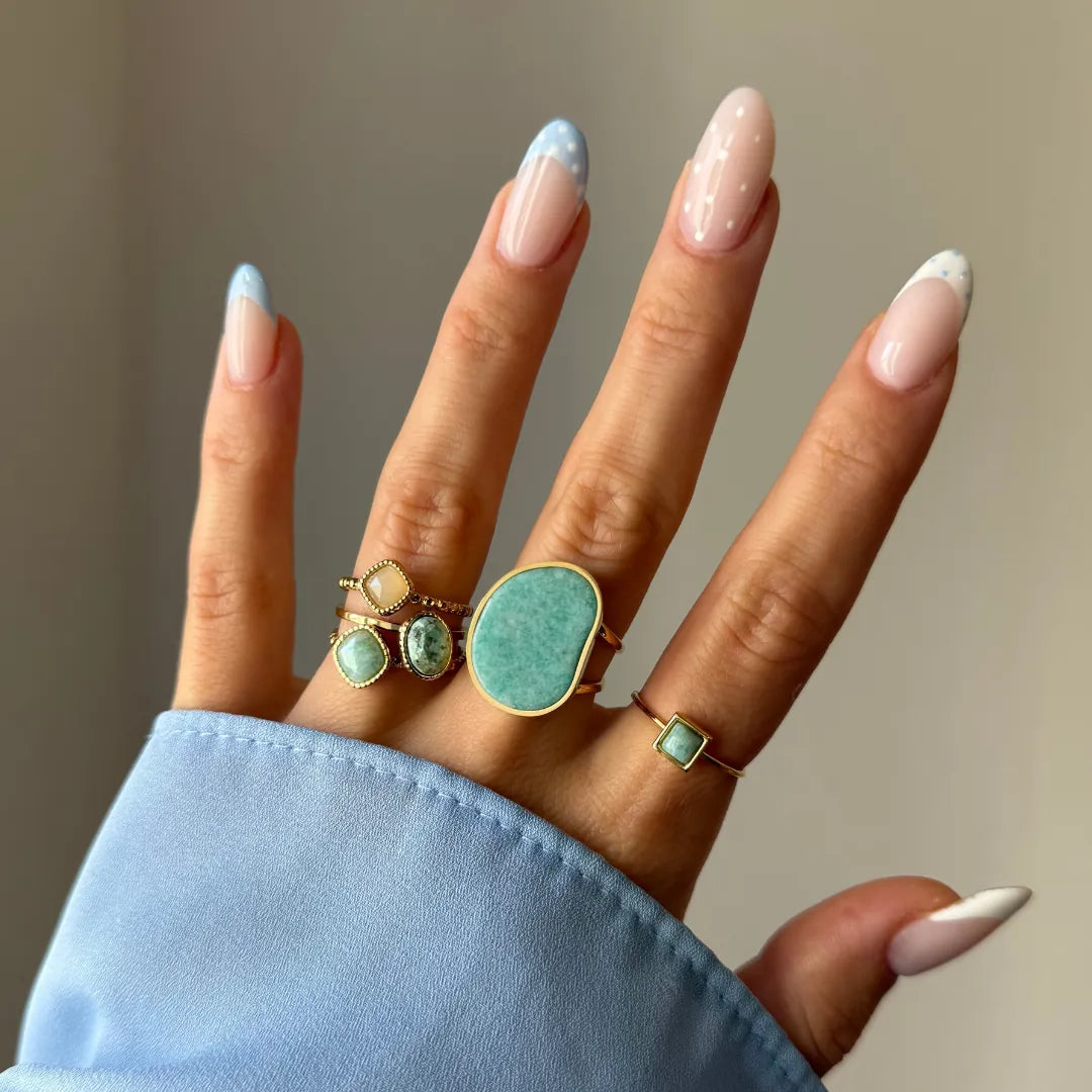 Main portant 5 bagues dorées aux pierres vert d'eau et jaune clair. Ongles manucurés avec motifs points blancs et bouts