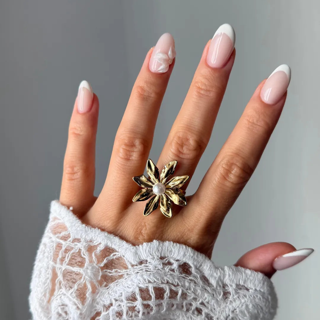 Bague fleur en acier inoxydable doré avec perle blanche, portée sur une main aux ongles manucurés et une manche en dentelle.