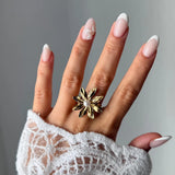 Bague fleur en acier inoxydable doré avec perle blanche, portée sur une main aux ongles manucurés et une manche en dentelle.