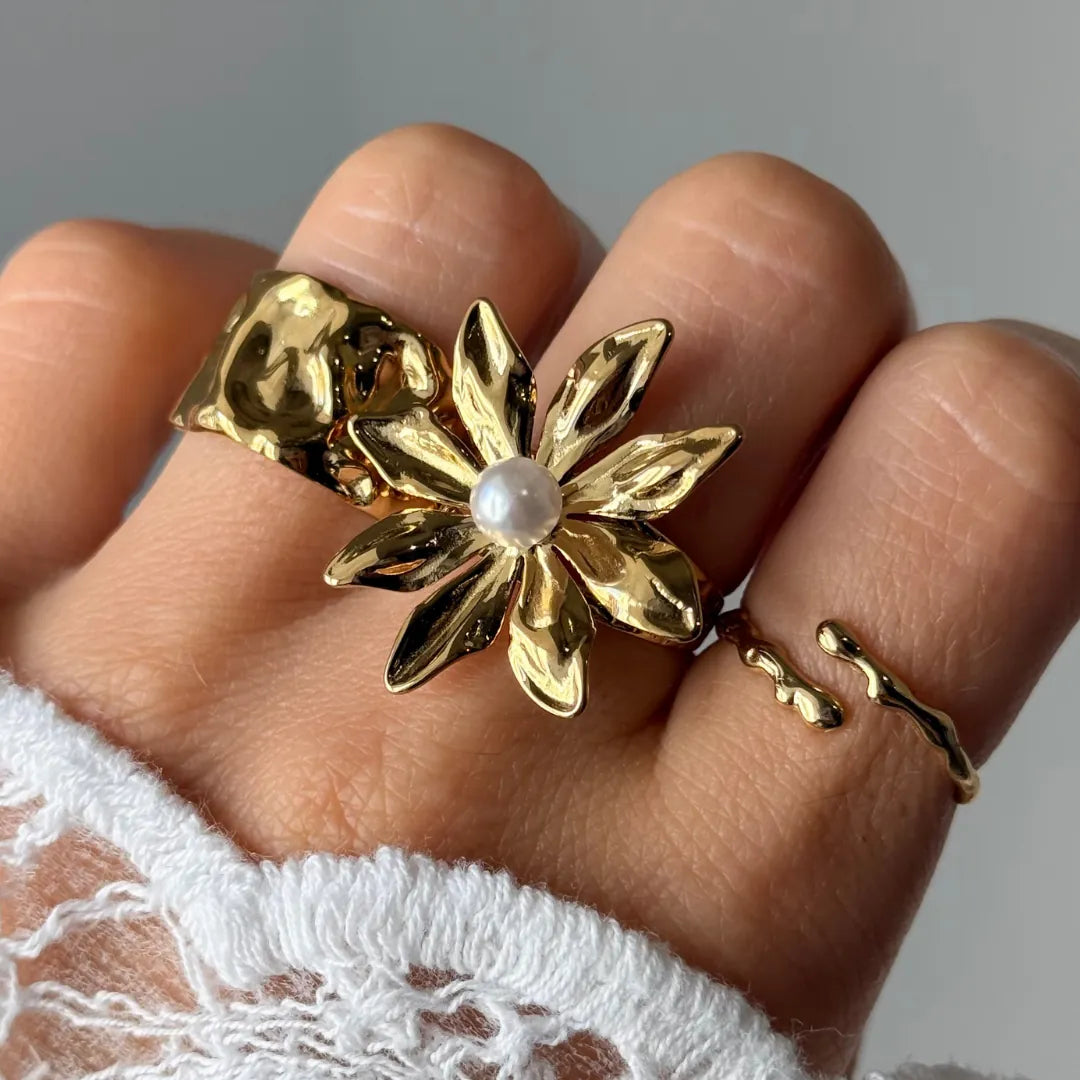 Main avec une bague fleur en acier inoxydable doré aux pétales texturés et une perle blanche, accompagnée de deux bagues