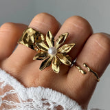 Main avec une bague fleur en acier inoxydable doré aux pétales texturés et une perle blanche, accompagnée de deux bagues