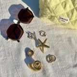 Bague étoile de mer texturée, d'autres bagues et un coquillage, tous en acier inoxydable doré, avec lunettes sur tissu blanc.