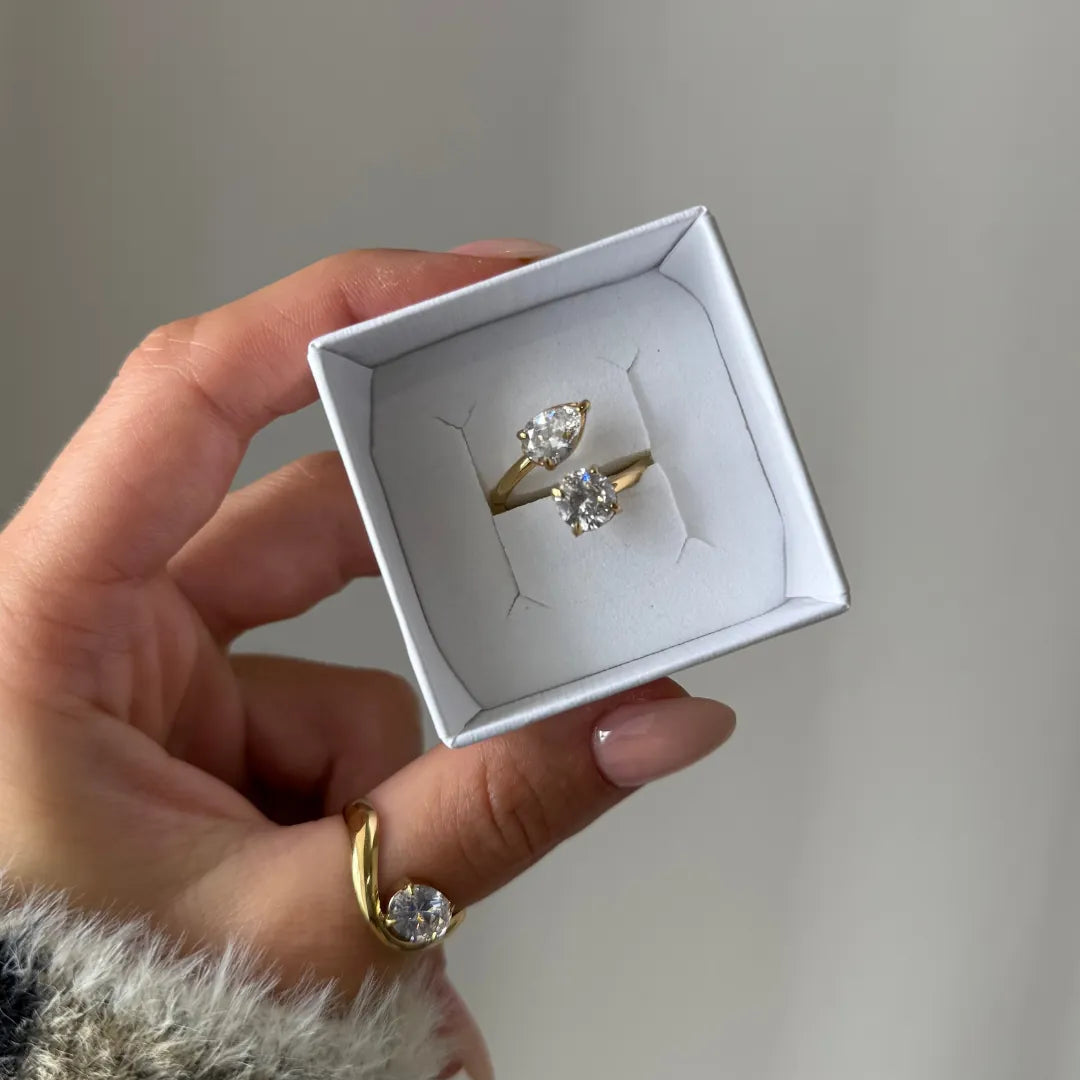 Bague toi et moi en acier inoxydable doré avec zirconium poire et rond, présentée dans un écrin blanc tenu par une main.