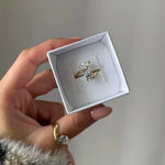 Bague toi et moi en acier inoxydable doré avec zirconium poire et rond, présentée dans un écrin blanc tenu par une main.