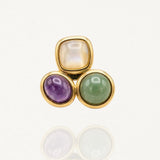 Bague dorée ornée de trois pierres cabochons géométriques (violet, vert et blanc nacré) vue de face sur fond blanc.