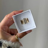 Main tenant une petite boîte blanche contenant une bague manchette multirangs en acier inoxydable doré, ornée de six pierres