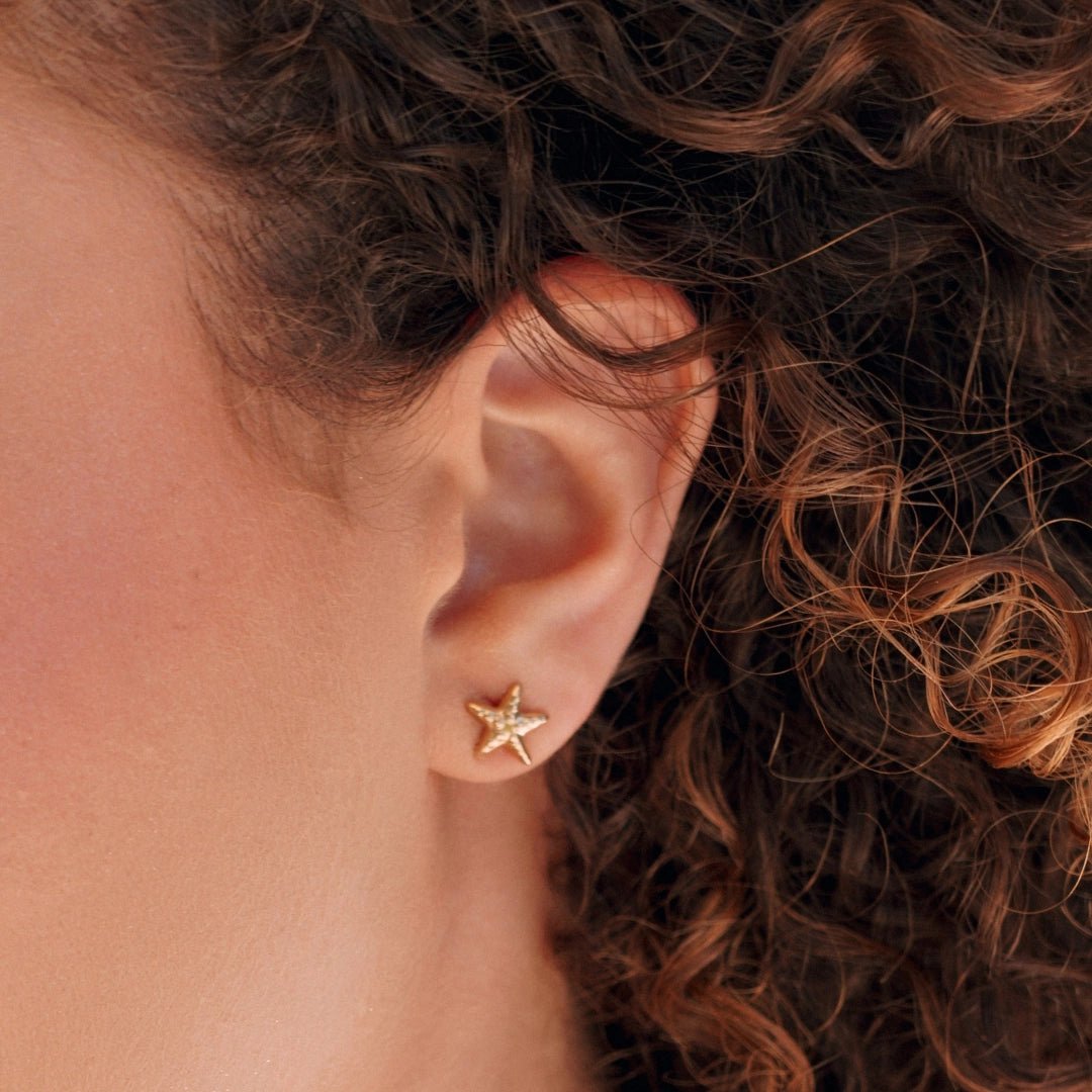 Boucles d'oreilles en acier inoxydable doré, forme étoile de mer texturée, portées sur le lobe d'oreille.