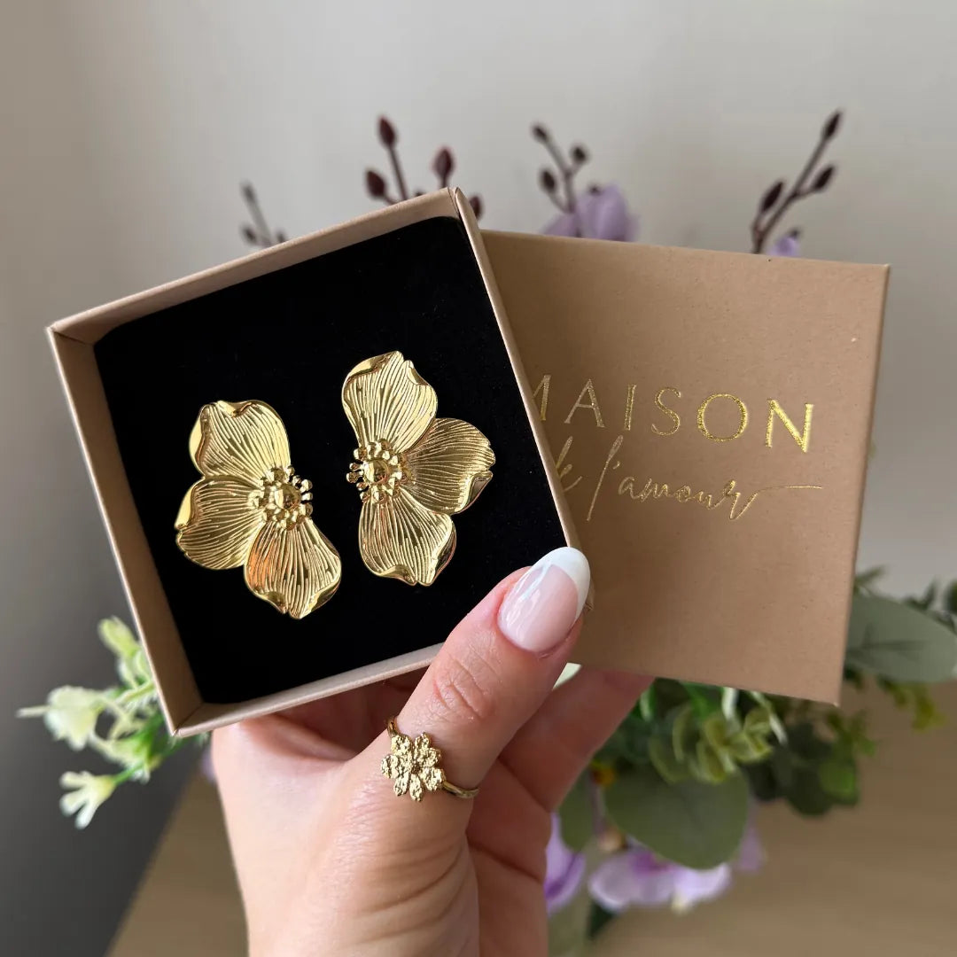 Deux boucles d'oreilles acier inoxydable dore en forme de fleurs, dans un écrin brun, tenu par une main portant une bague