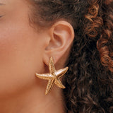 Boucles d'oreilles étoile de mer dorées en acier inoxydable avec texture détaillée, portées sur l'oreille d'une femme.