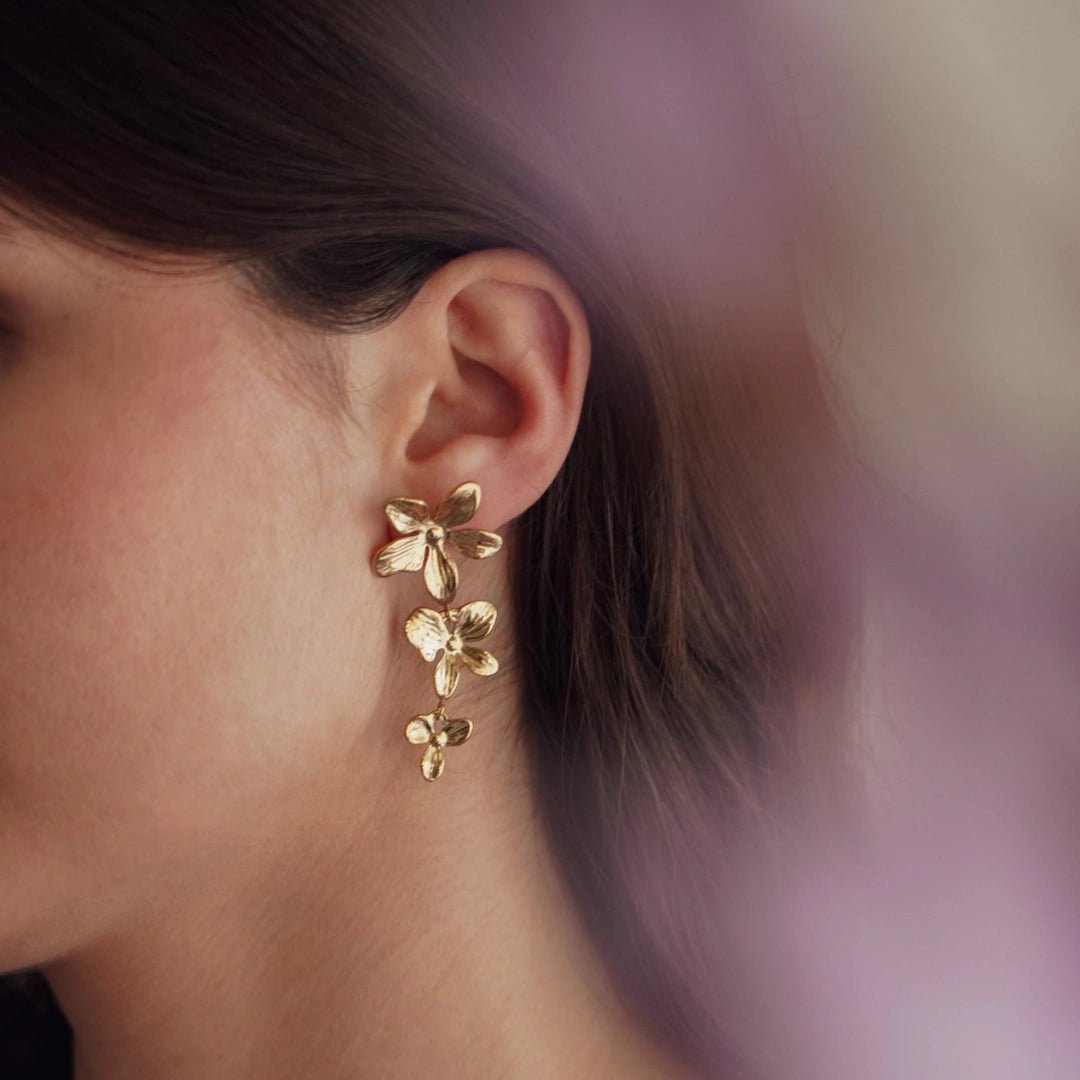 Boucles d'oreilles pendantes en acier inoxydable doré, à motif de trois fleurs, portées sur l'oreille.