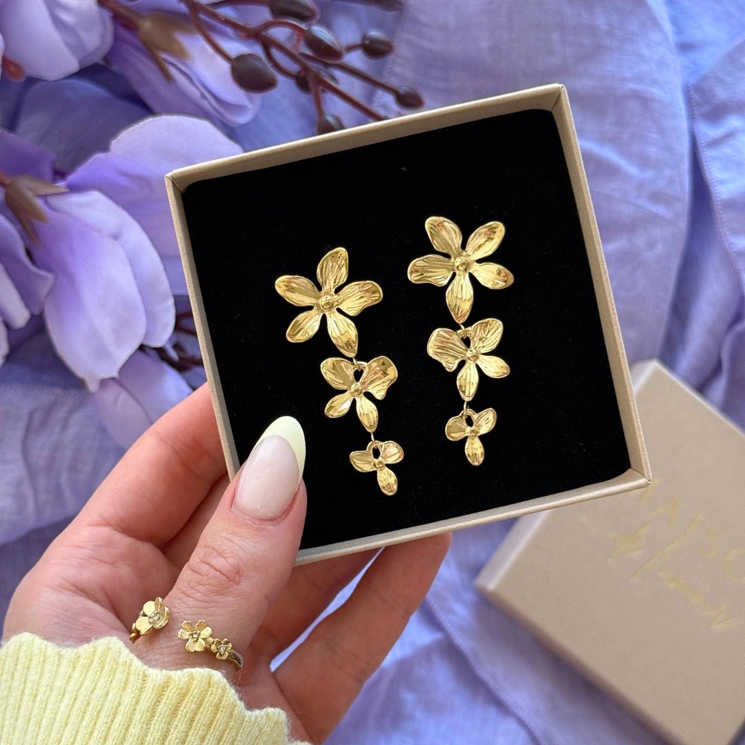 Boucles d'oreilles en acier inoxydable dorées, motif floral cascade, dans leur écrin tenu par une main.