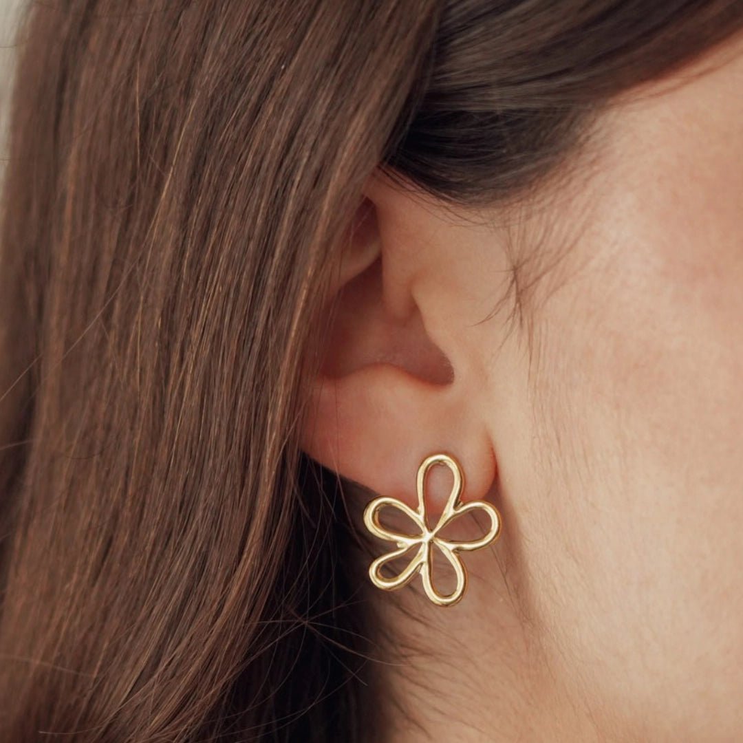 Boucles d'oreilles en acier inoxydable doré, motif fleur évidée, portées sur l'oreille d'une femme aux cheveux bruns.