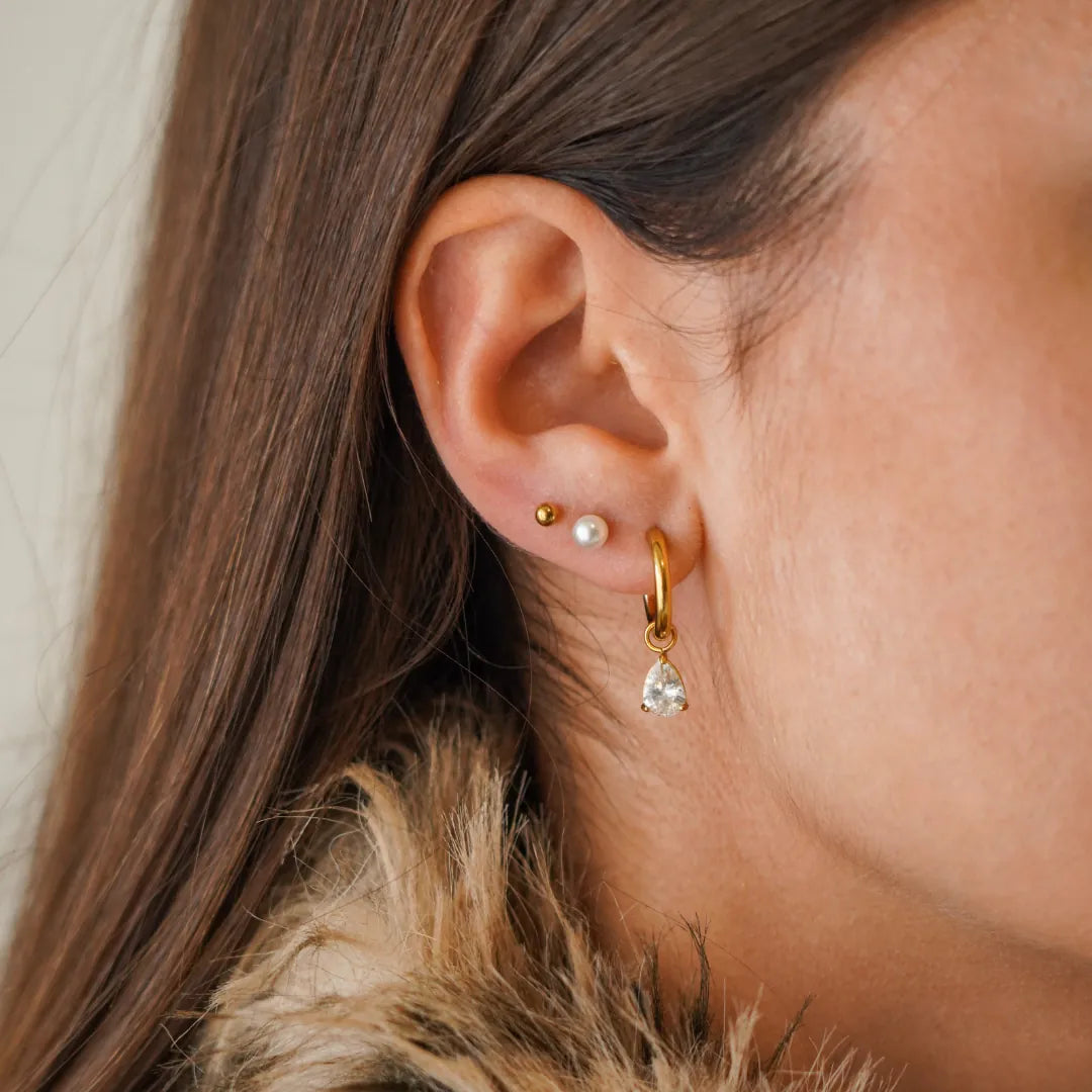 Gros plan sur une oreille avec trois boucles d'oreilles en acier inoxydable doré : une créole avec zirconium poire, une puce