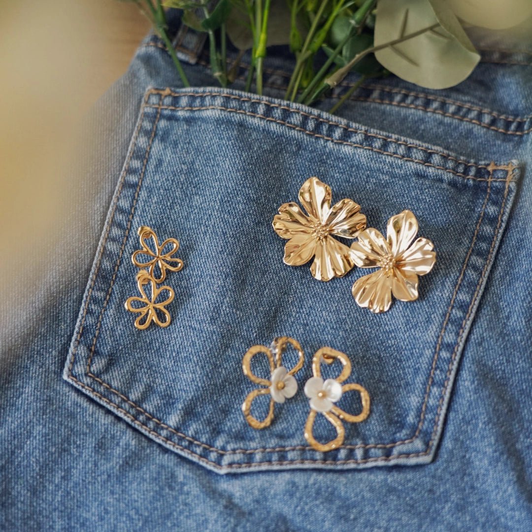 Trois paires de boucles d'oreilles en acier inoxydable doré, motifs fleurs variés, posées sur une poche de jean.