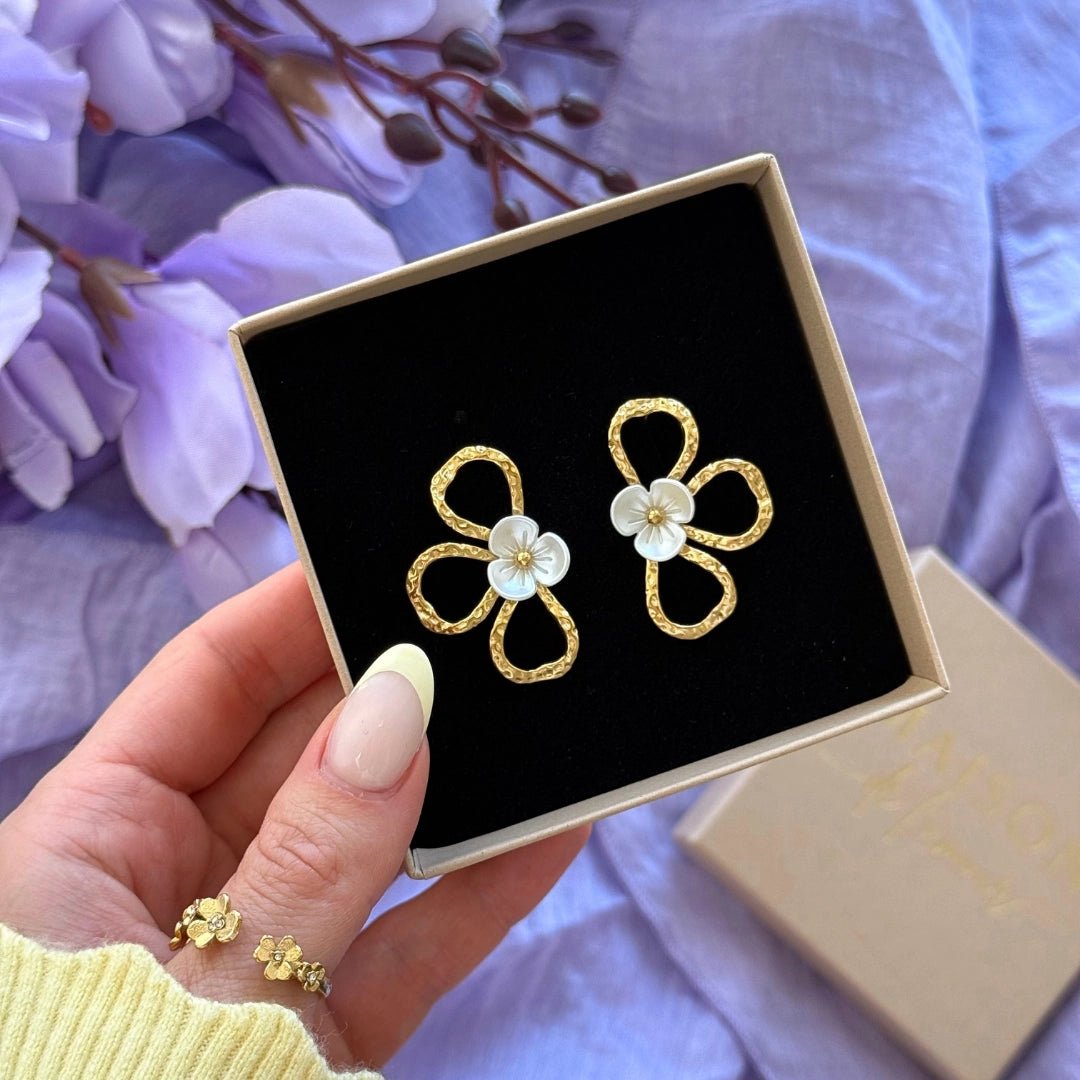 Boucles d'oreilles en acier inoxydable doré, motif trèfle martelé, avec petite fleur blanche centrale. Présentées dans un