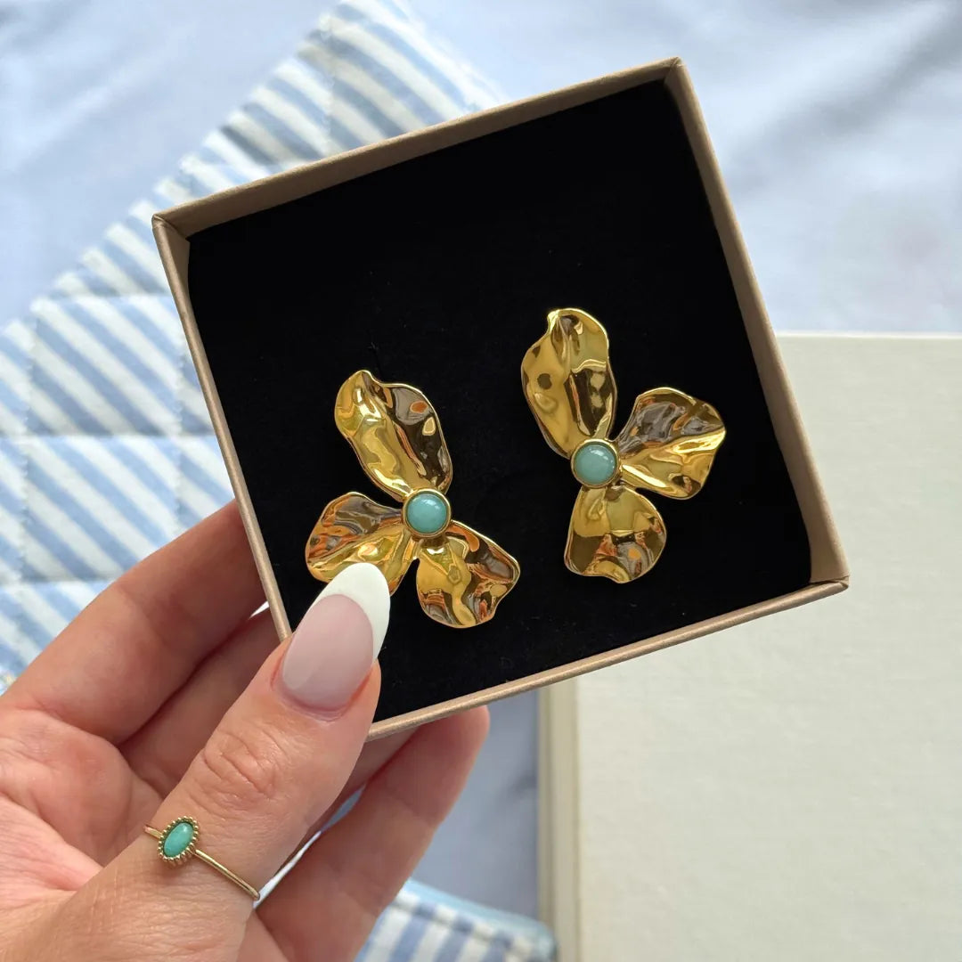 Boucles d'oreilles fleur en acier inoxydable doré texturé avec une pierre naturelle ronde bleu clair, dans un écrin noir.