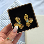Boucles d'oreilles fleur en acier inoxydable doré texturé avec une pierre naturelle ronde bleu clair, dans un écrin noir.