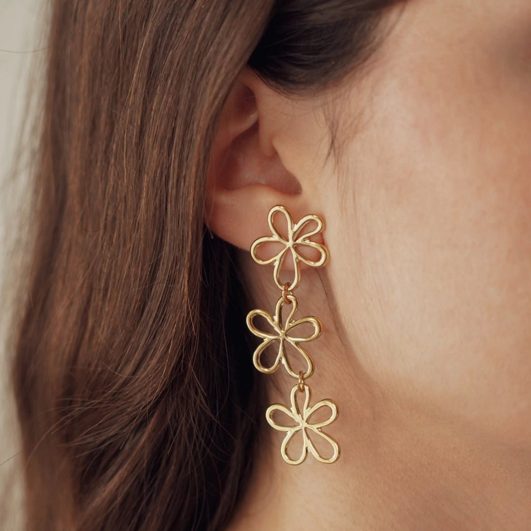 Boucles d'oreilles pendantes en acier inoxydable doré, motif 3 fleurs stylisées, portées sur une oreille.