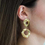 Boucle d'oreille pendante dorée martelée, composée de deux anneaux ouverts irréguliers, portée par femme aux cheveux bruns.