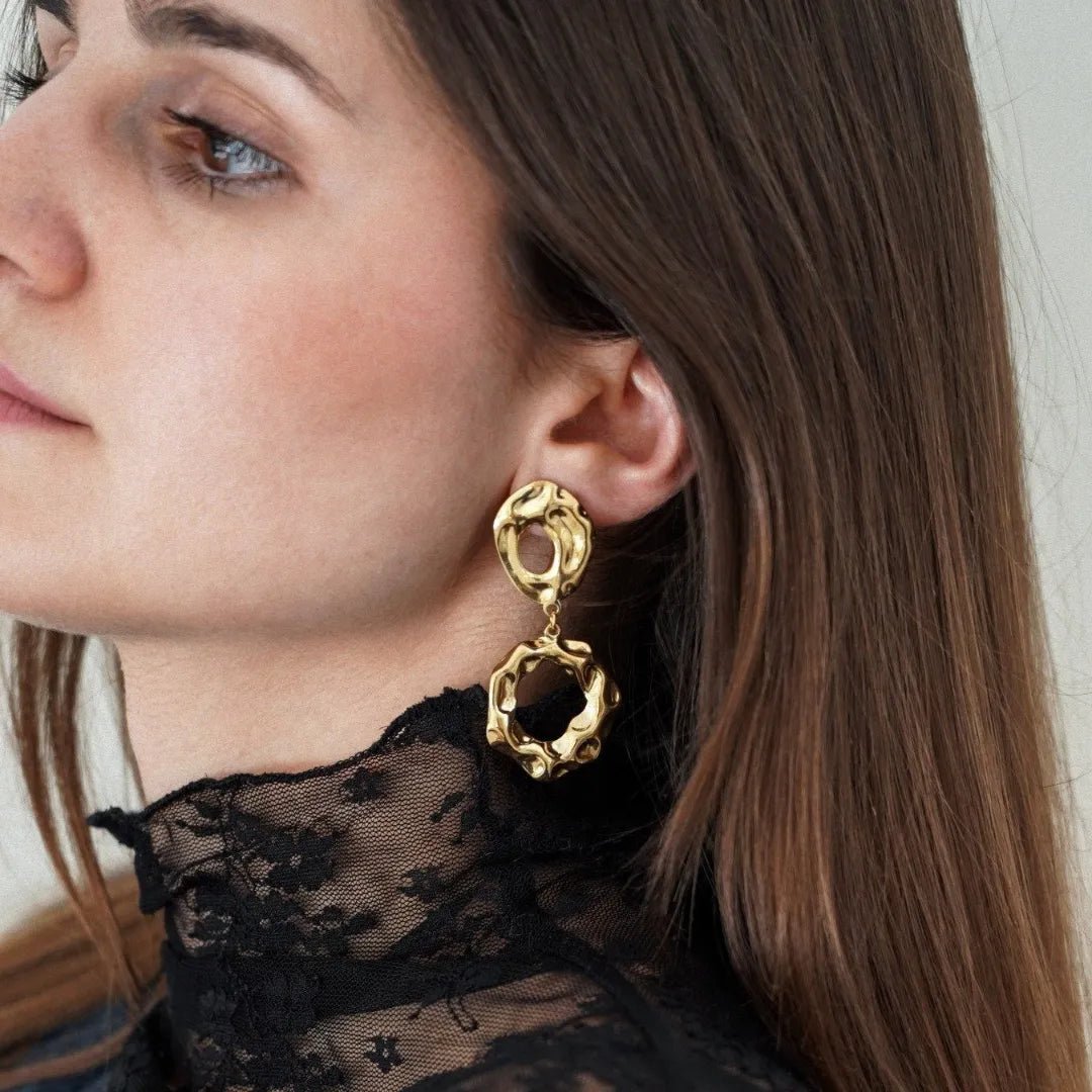 Profil de femme portant une boucle d'oreille pendante dorée texturée composée de deux anneaux irréguliers, et un col en dentelle noire.