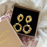 Paire de boucles d'oreilles pendantes dorées martelées, à deux éléments irréguliers avec trous, dans un écrin beige ouvert.