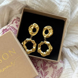 Paire de boucles d'oreilles pendantes dorées martelées, à deux éléments irréguliers avec trous, dans un écrin beige ouvert.