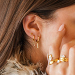 Sur une oreille, trois boucles d'oreilles en acier inoxydable doré et zirconium : une puce, une croix et une barrette.