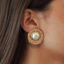 Boucles d'oreilles puces grand format style vintage avec large perle blanche et contour doré texturé portées à l'oreille.