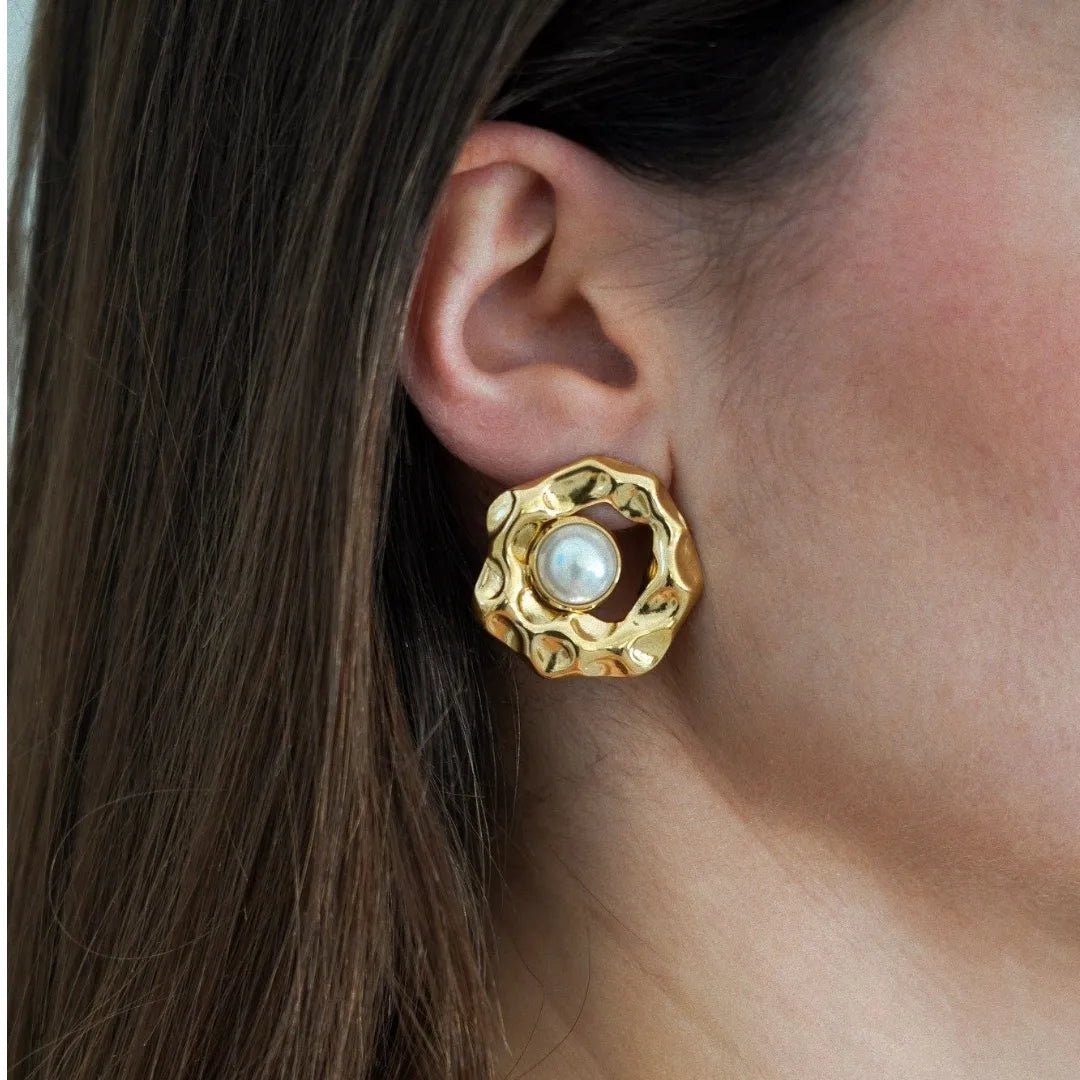 Gros plan sur une boucle d'oreille dorée, texture martelée, avec une perle blanche centrale, sur l'oreille d'une femme.