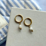 Deux boucles d'oreilles créoles en acier inoxydable doré, avec chacune un pendentif composé d'une bille blanche irrégulière