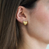 Boucle d'oreille clou cœur doré martelé portée sur l'oreille d'une femme aux cheveux bruns.