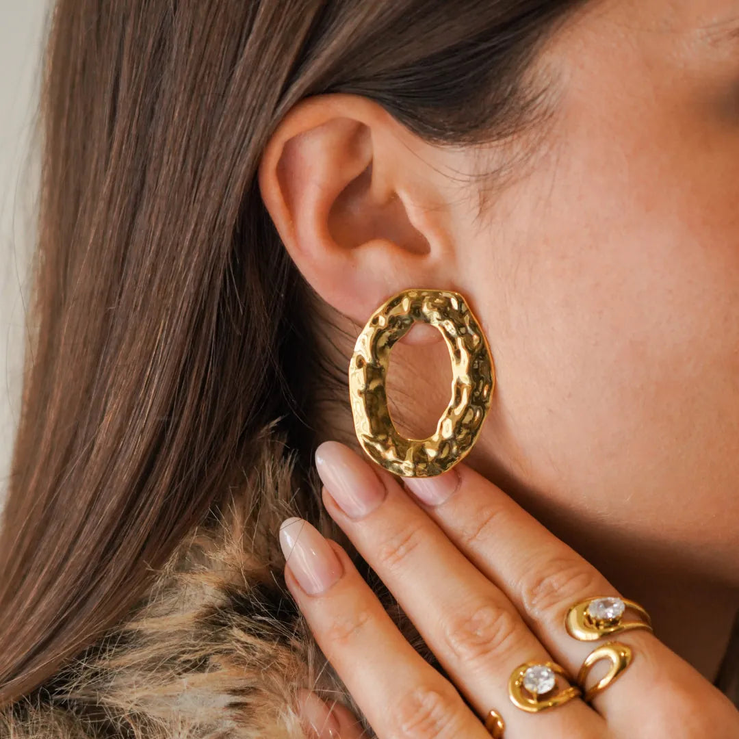 Une femme porte une boucle d'oreille ovale martelée en acier inoxydable doré, au relief organique. Sa main est près de son
