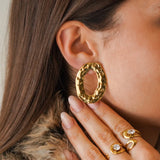 Une femme porte une boucle d'oreille ovale martelée en acier inoxydable doré, au relief organique. Sa main est près de son