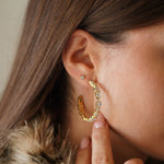 Créole tennis ovale en acier inoxydable doré et zircons, portée à l'oreille d'une femme. Sa main touche le lobe.