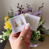 Main tenant une boîte avec boucles d'oreilles mini créoles en acier inoxydable doré, pendentifs fleurs et strass zirconium.