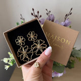 Paire de boucles d'oreilles pendantes à double fleur en acier inoxydable doré dans une boîte ouverte, tenue par une main.