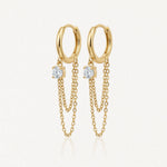 Paire de boucles d'oreilles créoles dorées avec chacune une pierre ronde brillante et deux chaînes fines pendantes.