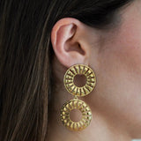 Boucle d'oreille pendante dorée, composée de deux cercles ajourés à motifs rayonnants, portée sur l'oreille droite d'une femme.