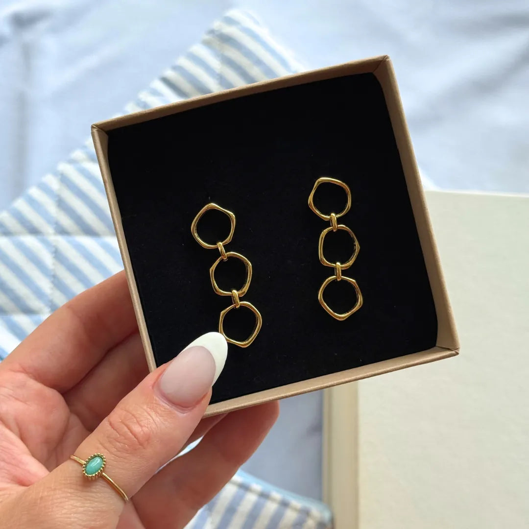 Paire de boucles d'oreilles en acier inoxydable doré, composées de trois maillons irréguliers, présentées dans un écrin noir.