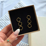 Paire de boucles d'oreilles en acier inoxydable doré, composées de trois maillons irréguliers, présentées dans un écrin noir.