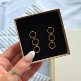 Paire de boucles d'oreilles en acier inoxydable doré, composées de trois maillons irréguliers, présentées dans un écrin noir.