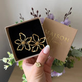 Main avec bague, tenant une boîte kraft "Maison de l'amour" contenant des boucles d'oreilles fleurs martelées en acier