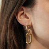 Gros plan sur la boucle d'oreille portée à l'oreille : focus sur la texture striée du maillon rectangulaire et la brillance du doré.